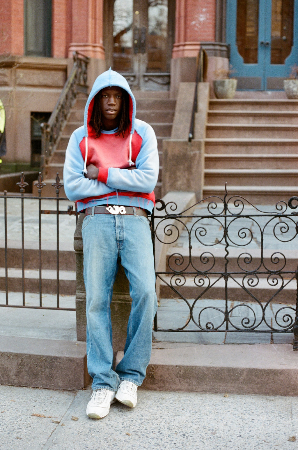 Person wearing a blue hoodie and jeans standing on a sidewalk with steps and a building in the background.